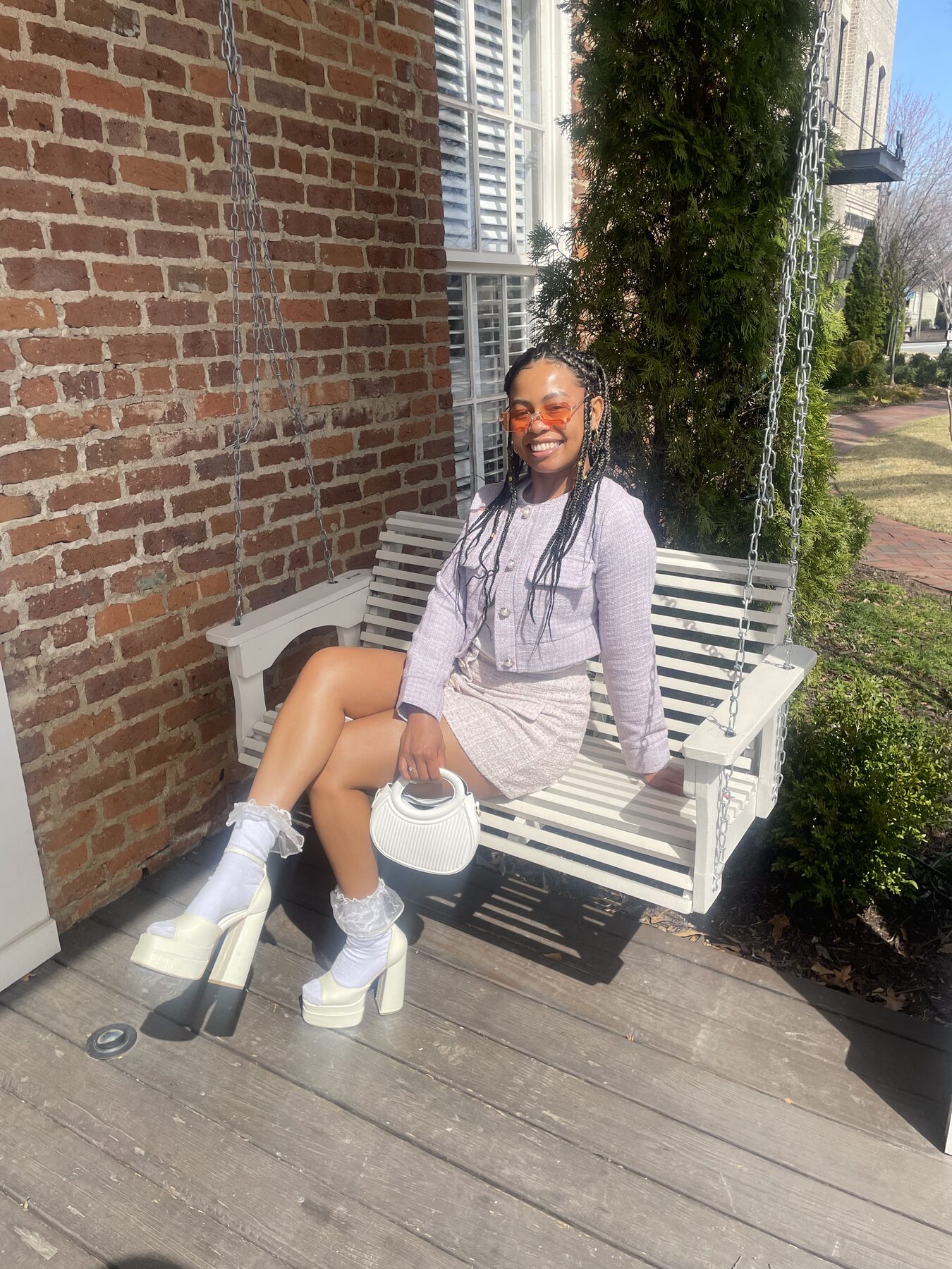 Tash Williamson seated on a white wooden porch bench swing, smiling broadly while holding a white quilted top-handle bag, wearing a lilac tweed jacket and matching mini skirt with white ruffle-trim socks and white platform boots, tinted orange sunglasses pushed up, long box braids, brick pavement and greenery behind her.