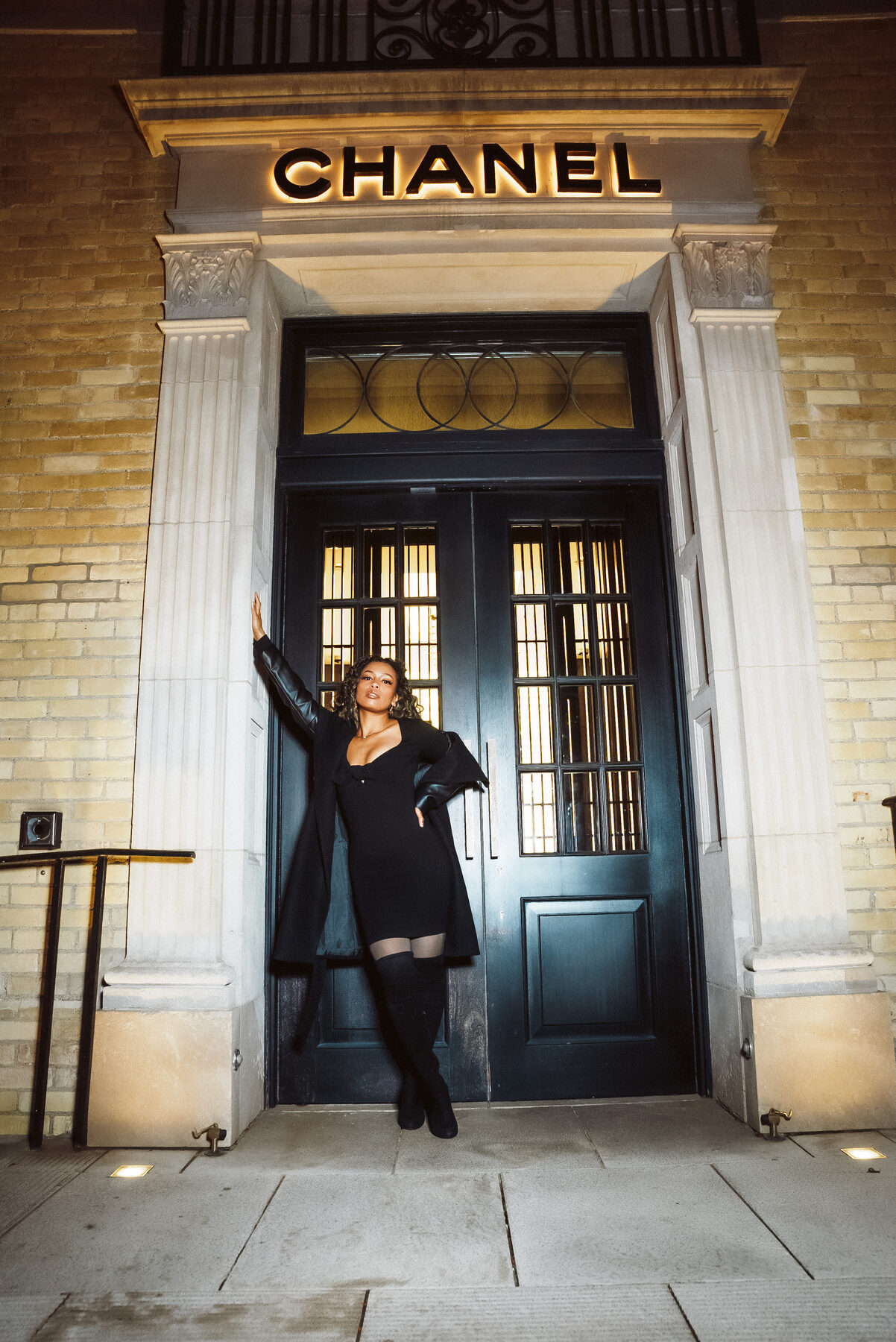 Tash Williamson in a full-length shot standing in the Chanel doorway, both arms raised overhead with hands in her hair, back arched and one leg crossed behind the other with her toe pointed against the door, eyes on the camera, black mini dress worn without the coat, sheer tights and thigh-high boots.