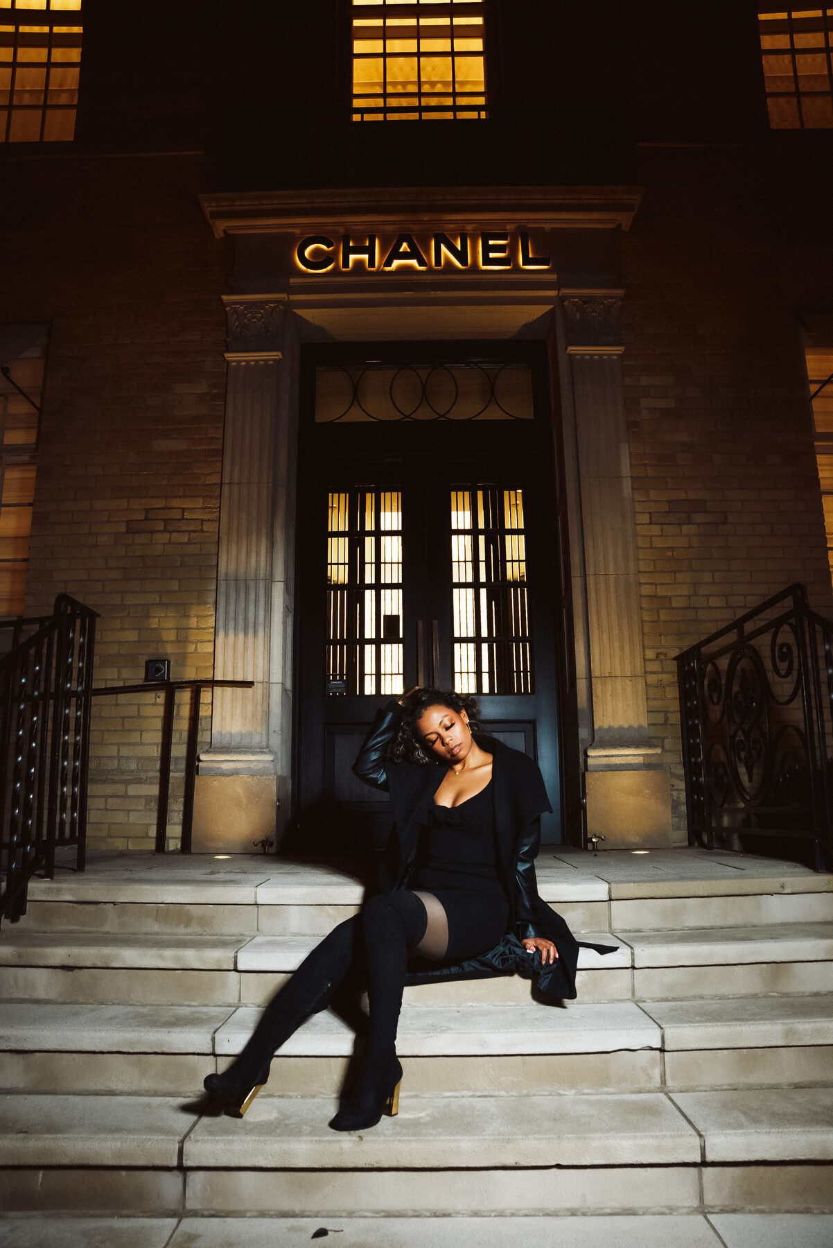 Tash Williamson in a full-length shot seated again on the Chanel storefront steps, head tipped back into her raised hand near her temple, eyes closed, legs bent and tucked to one side, black coat draped over her shoulders, thigh-high boots with gold heels catching the light.