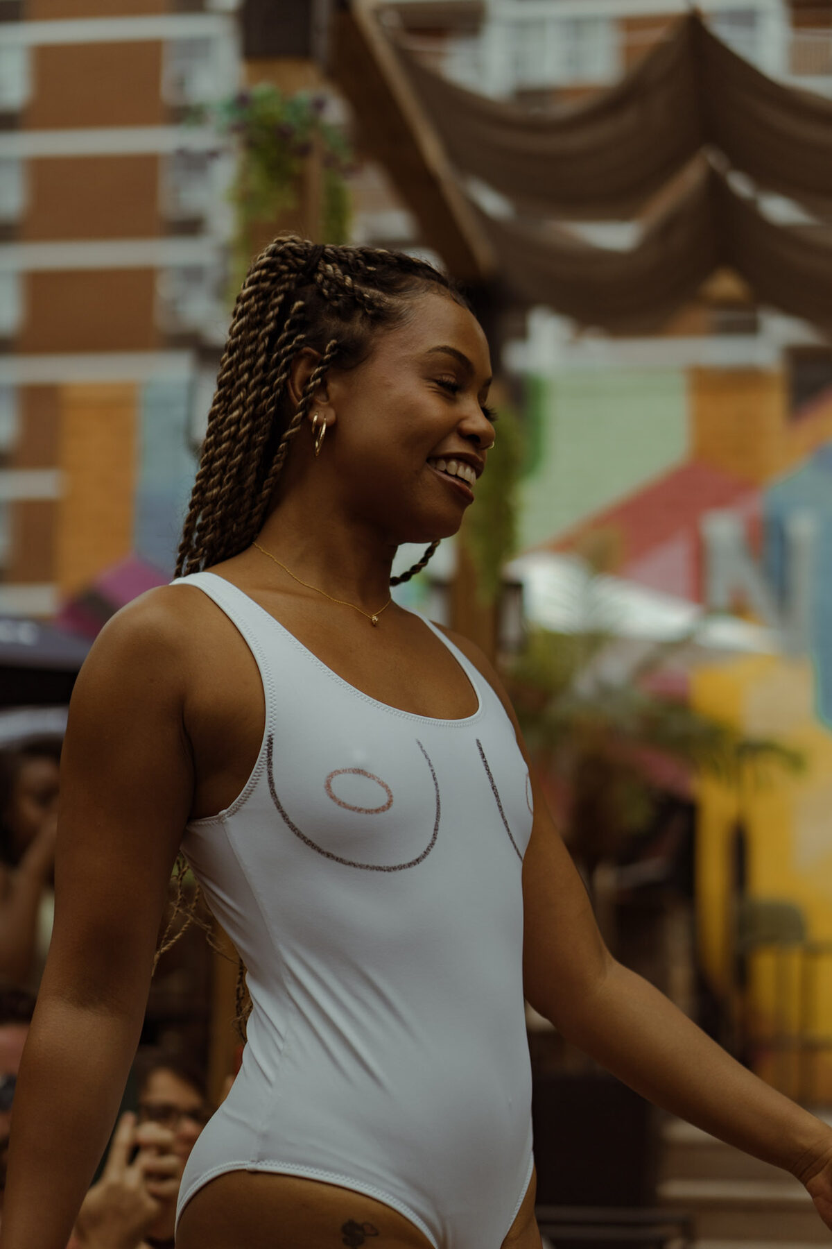 Tash Williamson caught in a candid moment outdoors, smiling and glancing off to the side while walking, wearing a fitted white tank bodysuit with a graphic heart outline printed across the chest, long braids over one shoulder, a colorful mural and market stalls blurred in the background.