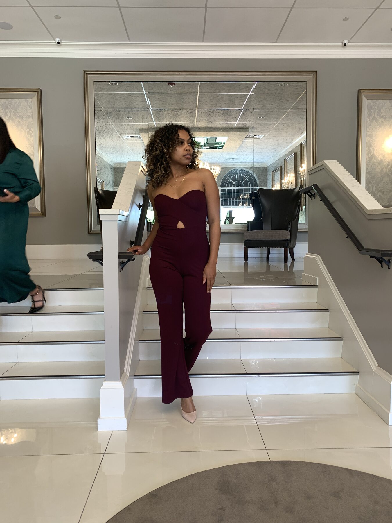 Tash Williamson in a near-identical staircase pose to a companion feature shot, leaning back against the same banister, but with her hand shifted up nearer her hairline and her gaze cast more directly toward the camera, chin tucked slightly lower, same burgundy cutout jumpsuit and loose curls.