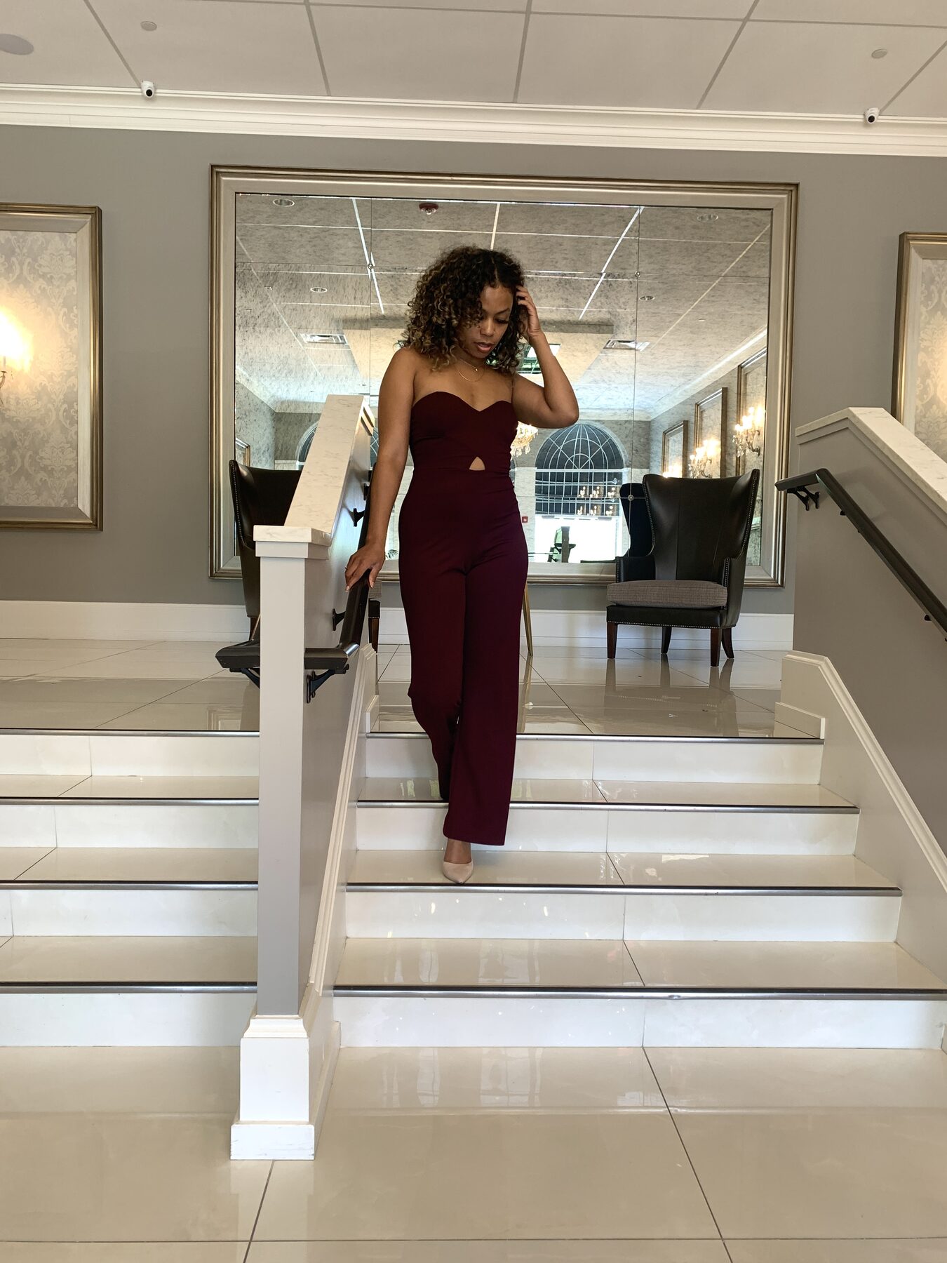 Tash Williamson in a full-length shot actually descending the staircase mid-step, one hand tucked into her curls and the other gripping the railing for balance, eyes cast down toward the stairs rather than at the camera, a mirrored hallway with dining chairs visible behind her, same burgundy jumpsuit as the staircase set.