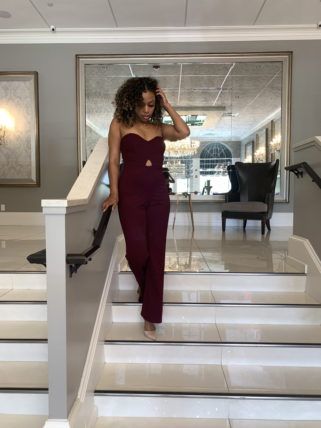 Tash Williamson in a full-length shot reclined further back against the staircase railing than the other frames in this set, head tilted back with eyes closed, one arm stretched behind her gripping the rail and the other hand near her hair, one leg extended forward as if mid-step, burgundy cutout jumpsuit.