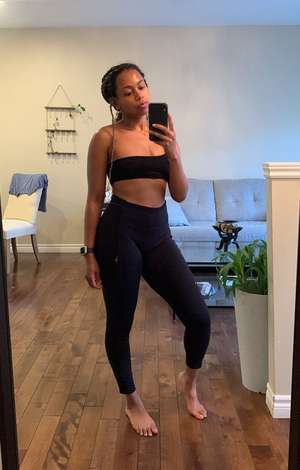 Tash Williamson in a full-length mirror selfie taken in a home living room, holding up her phone with one hand while her hair is styled in two long braids draped over one shoulder, wearing a black bandeau-style sports bra and high-waisted black leggings, barefoot on a wood floor, a smartwatch on her wrist, a gray couch, houseplant, and jewelry display visible in the background.