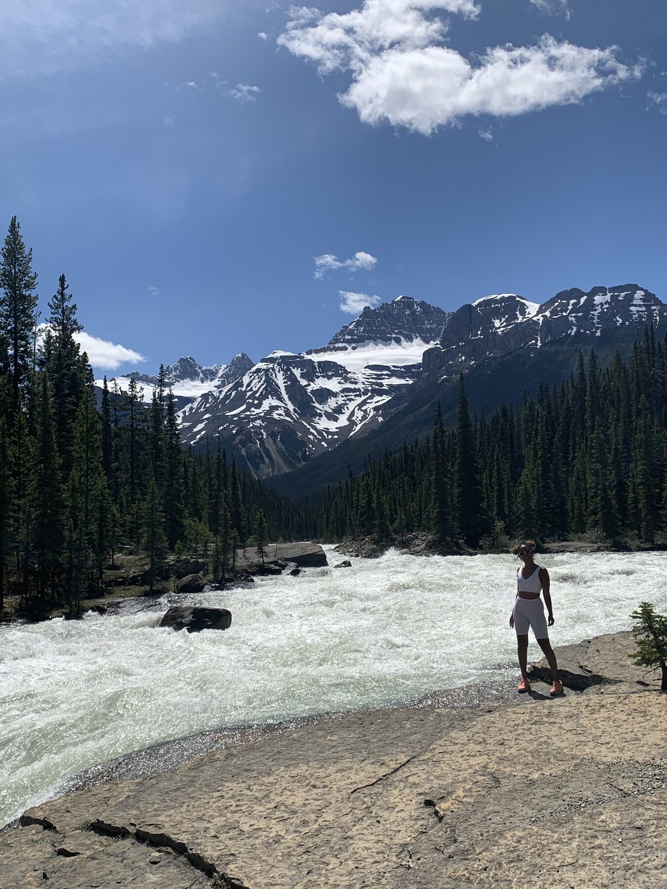 Tash Williamson walking along a rocky riverbank with a wide, fast-flowing glacial river and snow-capped mountain peaks behind her, wearing a white long-sleeve athletic top and matching leggings, one arm swinging as she strides forward — a candid, distant hiking snapshot rather than a posed shot.