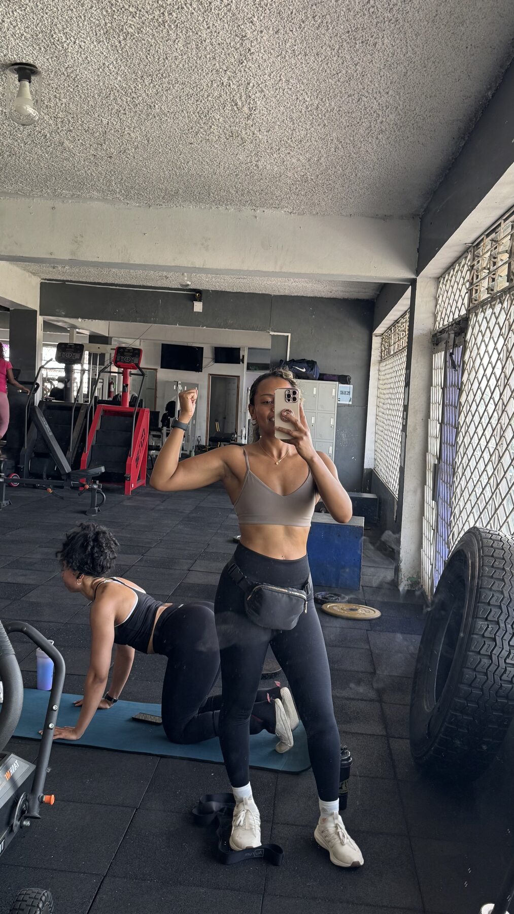 Tash Williamson in a gym mirror selfie, one arm raised flexing her bicep while the other holds up her phone, wearing a taupe sports bra, black leggings, and a black fanny pack worn at the waist, hair pulled back; the reflected gym shows workout machines, a large tire, and another woman exercising on a mat nearby.
