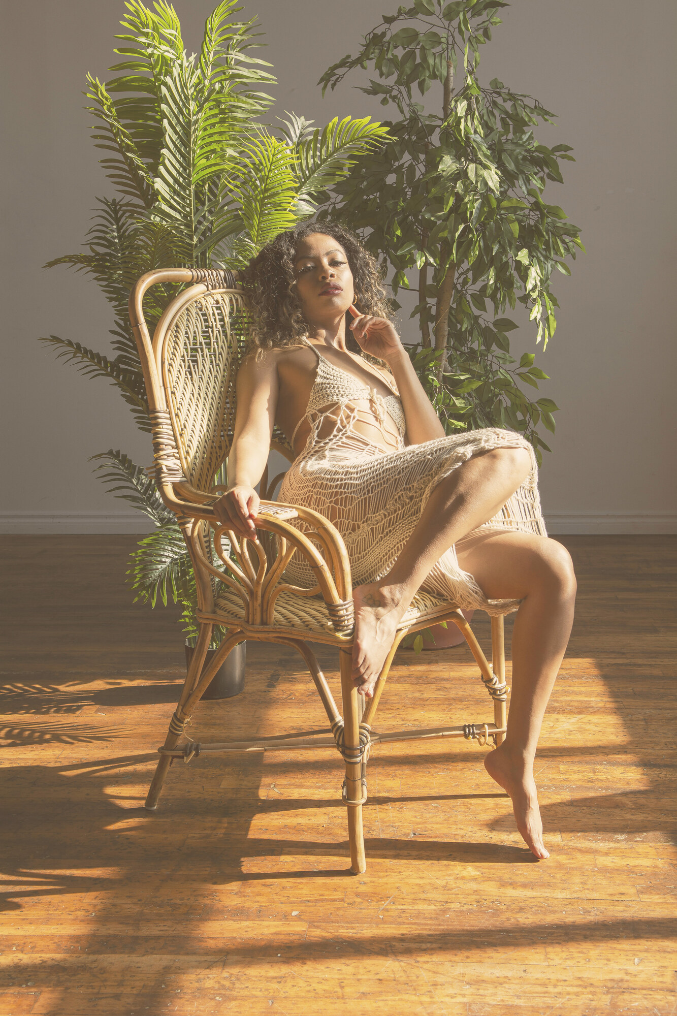 Tash Williamson in a full-length seated portrait in a rattan wingback chair, wearing a sheer cream crochet dress, one leg bent up onto the chair seat and the other extended to the floor, gazing directly at the camera with sunlit potted palms behind her.
